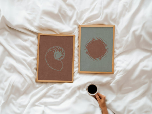 Two framed artworks on a bed with a hand holding a coffee cup.